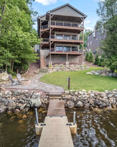 Waterfront house with multiple decks surrounded by trees, dock extending into water, and manicured lawn with stone landscaping.