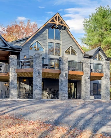 Modern two-story house with stone and siding exterior, multiple decks, and gable roof surrounded by fall foliage.