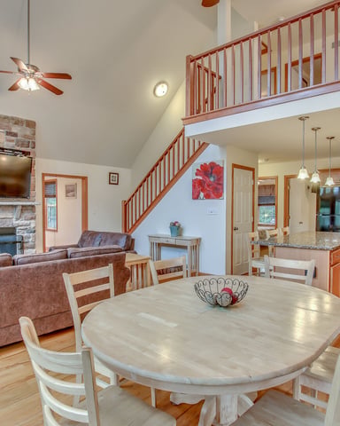 Open-plan living space with stone fireplace, dining table, brown sofa, kitchen, and loft overlooking main room