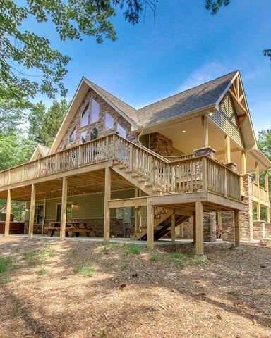 Two-story modern cabin with wraparound deck and stone chimney, surrounded by tall trees in wooded setting.