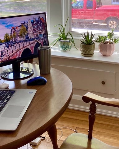Home office with round wooden desk, laptop, monitor displaying Amsterdam canal scene, potted plants on windowsill, wooden chair, and red truck visible outside.