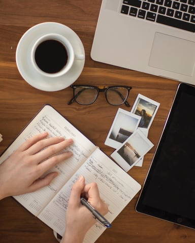 Overhead view of wooden desk with laptop, tablet, coffee cup, notebook, polaroid photos, glasses, and person's hand writing