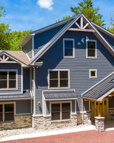 Modern two-story house with dark blue siding, stone foundation, two-car garage, and covered porch surrounded by tall trees