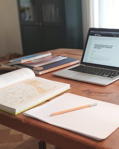 Wooden desk with laptop, open notebooks, sketches, coffee mug, and stacked books near a fireplace and window
