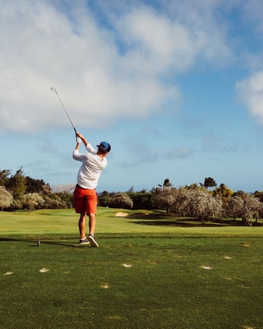 Golfer mid-swing on expansive fairway with olive trees and blue sky
