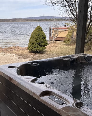 Hot tub on sandy beach overlooking calm lake with bare trees and distant shoreline