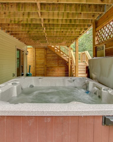Hot tub on covered wooden deck with stairs and lattice roof, adjacent to house with green siding