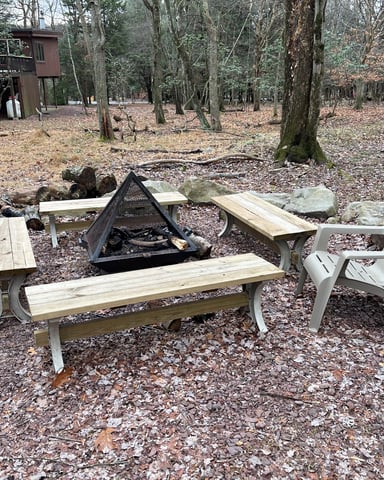 Wooded campfire area with wooden benches, metal fire pit, and Adirondack chairs surrounded by tall trees and cabins in background