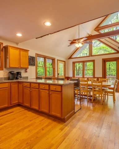 Open-plan kitchen and dining area with wood cabinets, island, hardwood floors, and vaulted ceiling with exposed beams overlooking forest.