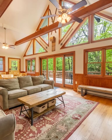 Spacious living room with exposed wood beams, vaulted ceiling, multiple windows overlooking forest, and wood-framed glass doors to deck