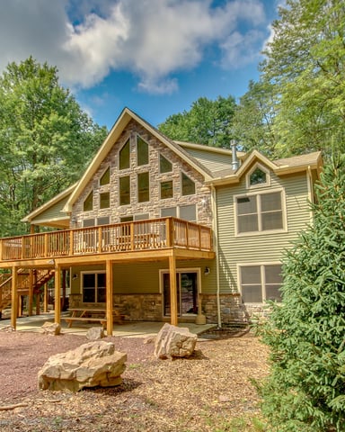 Modern house with stone and siding exterior, wooden deck, and fire pit surrounded by picnic tables and tall pine trees.