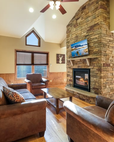 Open-plan living room with vaulted ceiling, stone fireplace, brown leather seating, and large windows overlooking water.