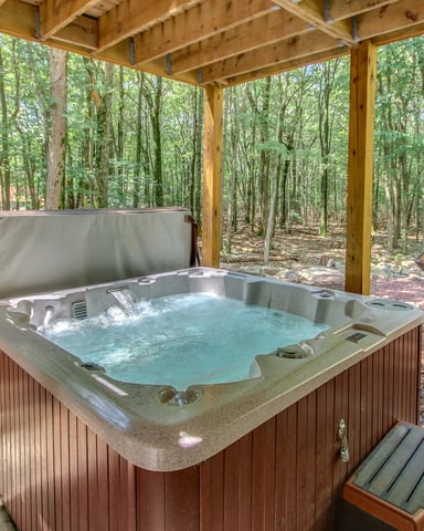 Hot tub with steaming water under wooden pergola in forest clearing, surrounded by tall trees