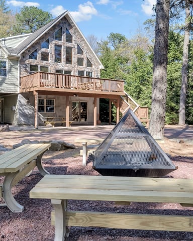Modern house with stone and siding exterior, wooden deck, and fire pit surrounded by picnic tables and tall pine trees.