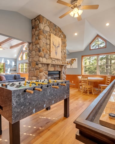 Game room with foosball table, shuffleboard, stone fireplace, and wood-paneled walls with large windows overlooking trees