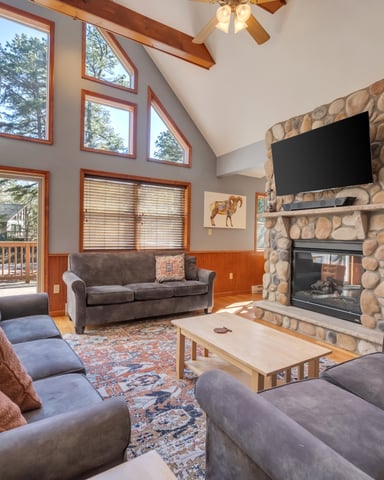 Spacious living room with stone fireplace, TV, gray sofas, wood beams, and large windows overlooking trees and deck
