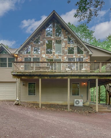 Two-story stone and siding house with peaked roof, upper deck, garage, and covered porch surrounded by tall trees