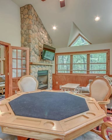 Game room with octagonal poker table, wood-trimmed windows, stone fireplace, and open seating area with forest views