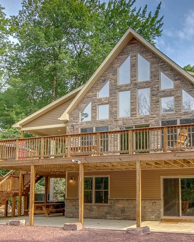 Two-story stone and wood house with large deck and covered porch, surrounded by tall trees