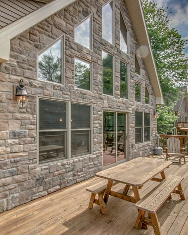 Wooden deck with picnic table and Adirondack chairs facing stone house with large windows and wooded yard