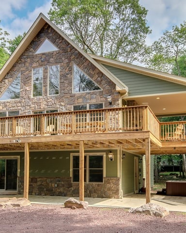 Modern stone and wood cabin with peaked roof, wraparound deck, and covered patio nestled among tall trees.