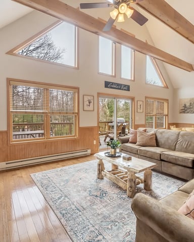 Vaulted living room with exposed wood beams, stone fireplace, multiple windows, ceiling fans, and upholstered seating overlooking a deck.