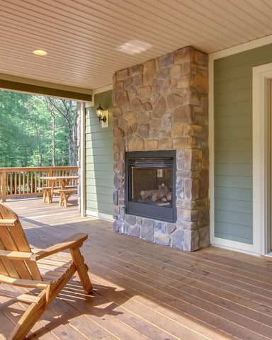 Covered deck with wooden Adirondack chairs, stone fireplace, and glass doors overlooking forested landscape
