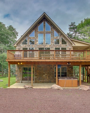 Modern A-frame cabin with stone exterior, large windows, wraparound deck, and hot tub surrounded by tall pine trees