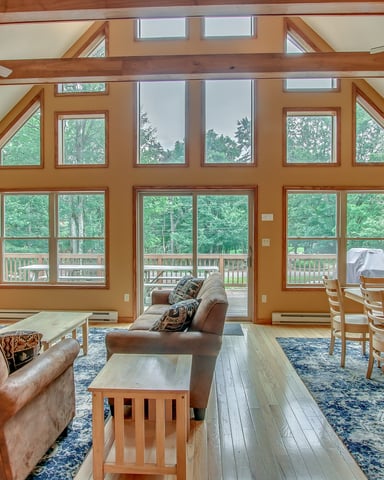 Open-plan living room with exposed wood beams, leather seating, multiple windows overlooking forest, and dining area with wooden chairs.
