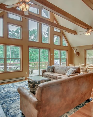 Spacious vaulted living room with exposed wood beams, leather seating, stone fireplace, and expansive windows overlooking wooded lake views.