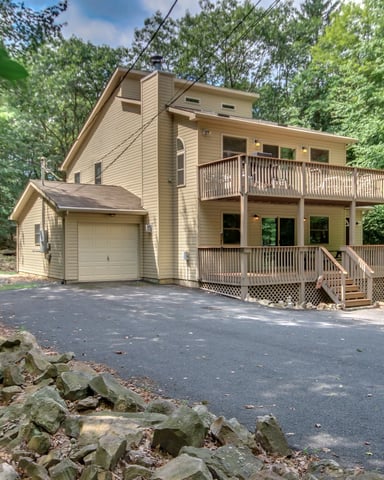 Two-story tan house with wraparound decks nestled among dense green trees, driveway with attached garage in foreground.
