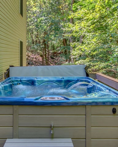 Blue hot tub on wooden deck with gray cover, surrounded by dense green forest and tan house siding