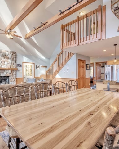 Open-plan dining and living area with vaulted ceilings, wooden beams, stone fireplace, and kitchen visible in background