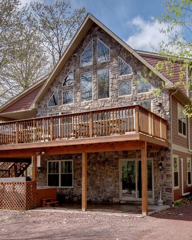 Two-story stone and wood house with large deck and covered porch, surrounded by bare and budding trees