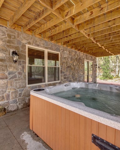 Covered patio with hot tub, stone wall, wooden beams, and wooded surroundings