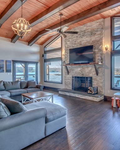 Open-plan living room with exposed wood beams, stone fireplace with TV, gray sectional sofa, and lake views through large windows