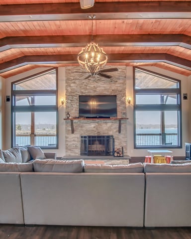 Open-plan living room with exposed wood beams, brick fireplace, large windows overlooking water, and gray sectional sofa.