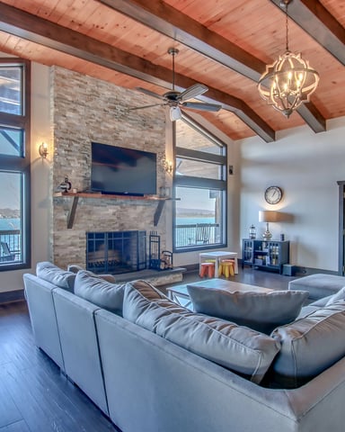 Open-plan living room with gray sectional sofa, exposed wood ceiling beams, brick fireplace with TV, and large windows overlooking a lake.