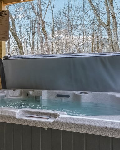 Hot tub with turquoise water under wooden pergola, bare trees visible in background forest setting