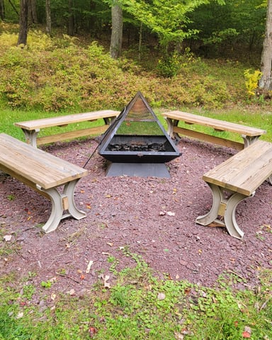 Outdoor fire pit surrounded by four wooden benches in a wooded clearing with green grass and trees.