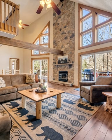 Open-plan living room with stone fireplace, vaulted ceilings, leather seating, and large windows overlooking wooded landscape.