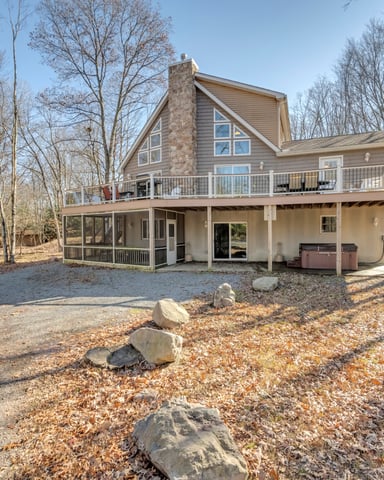 Two-story house with stone chimney and wraparound deck surrounded by bare trees and gravel driveway