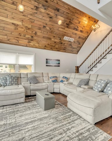 Open-plan living room with cream sectional sofa, wooden plank ceiling, staircase, and glass doors opening to deck