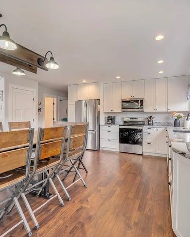 Open-plan kitchen and dining area with white cabinetry, stainless steel appliances, wood dining table, and potted plants on shelving.