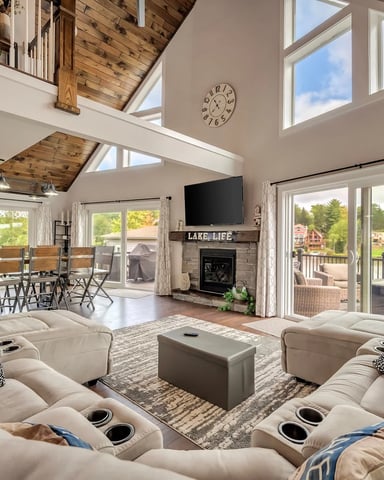 Open-plan lake house living room with vaulted wood ceiling, cream sectional seating, fireplace, and expansive windows overlooking water and trees.