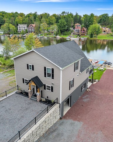 Waterfront house with gray siding and dark roof on river, surrounded by trees and neighboring homes across the water.