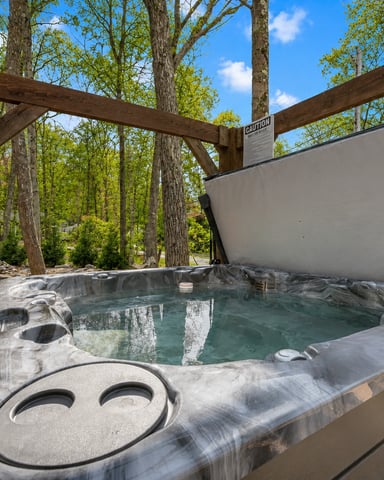 Hot tub with retractable shade cover under wooden pergola surrounded by tall trees and clear sky