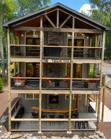 Three-story treehouse-style cabin with stone chimney, multiple decks, and black metal railings surrounded by tall trees