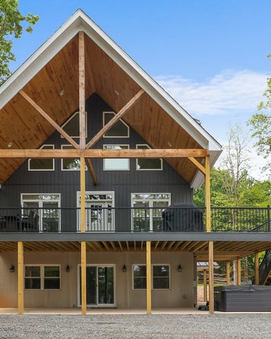 Modern barn-style house with black upper level, wooden beams, and covered deck surrounded by tall trees
