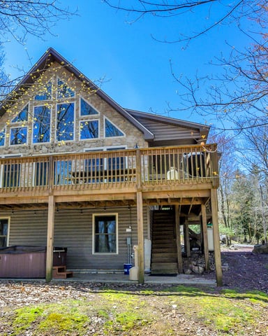 Two-story wooden cabin with large upper-level windows, wraparound deck, and hot tub, surrounded by bare trees